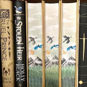 Op de afbeelding: Een set van vier hardcover boeken met gouden accenten en geïllustreerde omslagen met een blauwe vogel, draken en bergen. De boeken zijn getiteld "The Stolen Heir" door Holly Black.