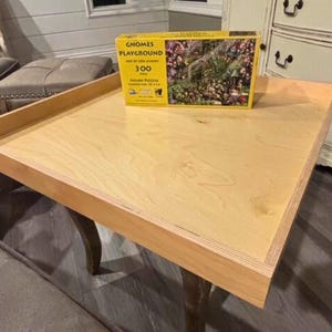 May include: A light wood-toned puzzle table with a raised edge, holding a puzzle box labelled "Gnomes Playground." The table has curved legs and is set in a room with neutral tones.