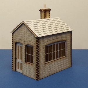 Puede incluir: Un pequeño modelo de madera de un edificio de ladrillo con un techo blanco y una chimenea. El edificio tiene una puerta y una ventana.