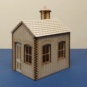 Pode incluir: Um pequeno modelo de madeira de um edifício de tijolo com um telhado branco e uma chaminé. O edifício tem duas janelas e uma porta.