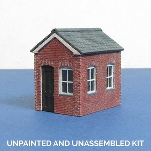 Pode incluir: Um modelo em miniatura de um edifício de tijolo com telhado cinza escuro, caixilhos de janelas brancos e uma porta preta. O texto "UNPAINTED AND UNASSEMBLED KIT" está na parte inferior. O modelo está sobre uma superfície azul.