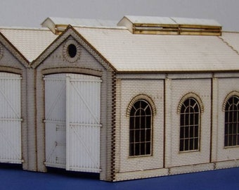 May include: A model train building made of wood with a white brick facade, two large doors, and several windows. The building has a brown roof and is painted in a light brown color.