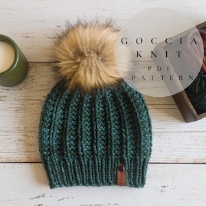 May include: A green knitted beanie with a brown faux fur pom pom. The beanie has a ribbed texture and a brown leather tag. The text "GOCCIA KNIT PDF PATTERN" is visible in a circle.