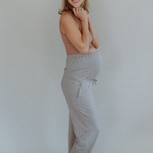 May include: A woman wearing a light brown tank top and light gray maternity pants. The pants have a drawstring and a wrap-around design. The woman is smiling and has her hands near her face.