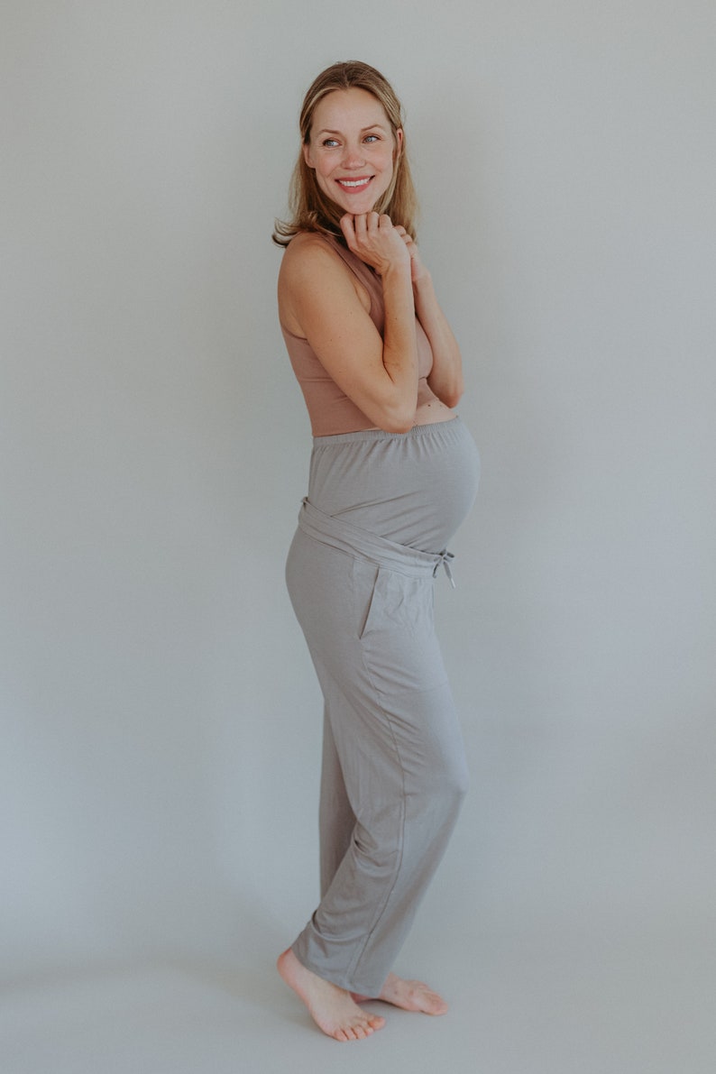 May include: A woman wearing a light brown tank top and light gray maternity pants. The pants have a drawstring and a wrap-around design. The woman is smiling and has her hands near her face.