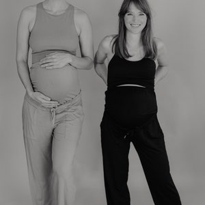 May include: Two pregnant people wearing maternity activewear. One wears a peach tank top and matching pants. The other wears a black tank top and black pants. Both are smiling and standing in front of a gray background.