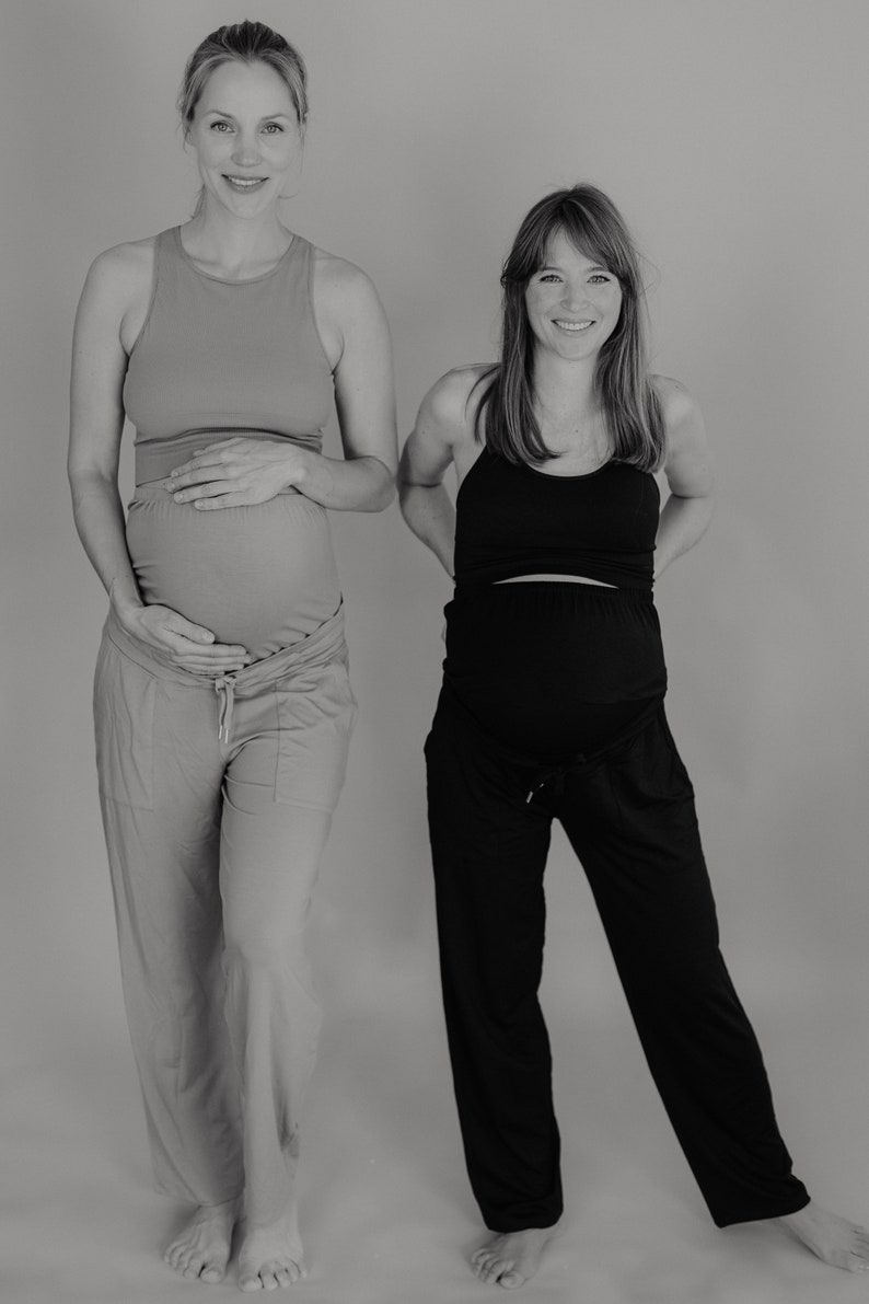 May include: Two pregnant people wearing maternity activewear. One wears a peach tank top and matching pants. The other wears a black tank top and black pants. Both are smiling and standing in front of a gray background.
