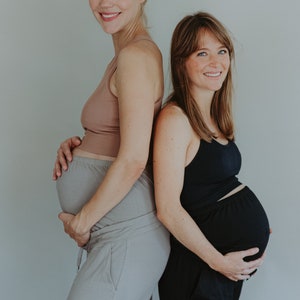 May include: Two pregnant individuals stand back-to-back, smiling at the camera. One wears a light brown tank top and gray pants, the other a black tank top and black pants. Both are holding their bellies, posing against a plain gray background.