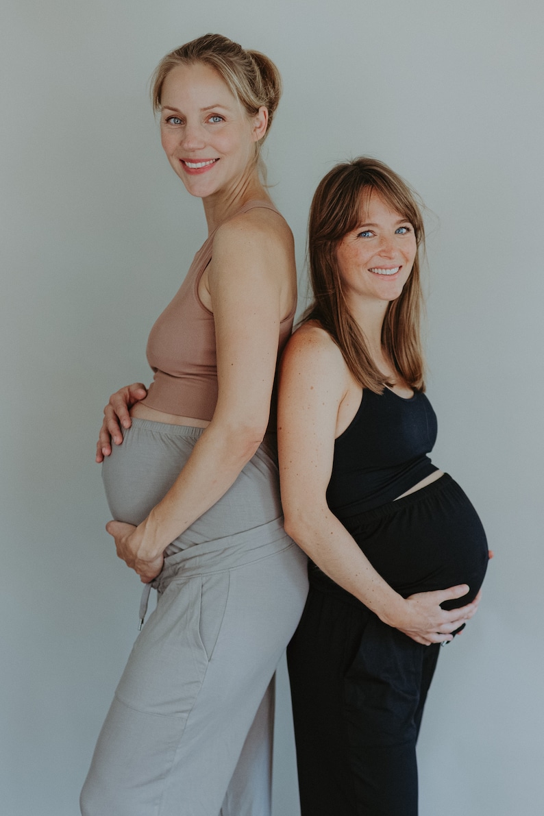 May include: Two pregnant individuals stand back-to-back, smiling at the camera. One wears a light brown tank top and gray pants, the other a black tank top and black pants. Both are holding their bellies, posing against a plain gray background.