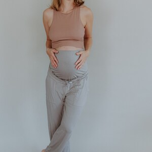 May include: A woman wearing a light brown tank top and gray maternity pants. The pants have a drawstring waist and pockets. The woman is smiling and holding her belly.
