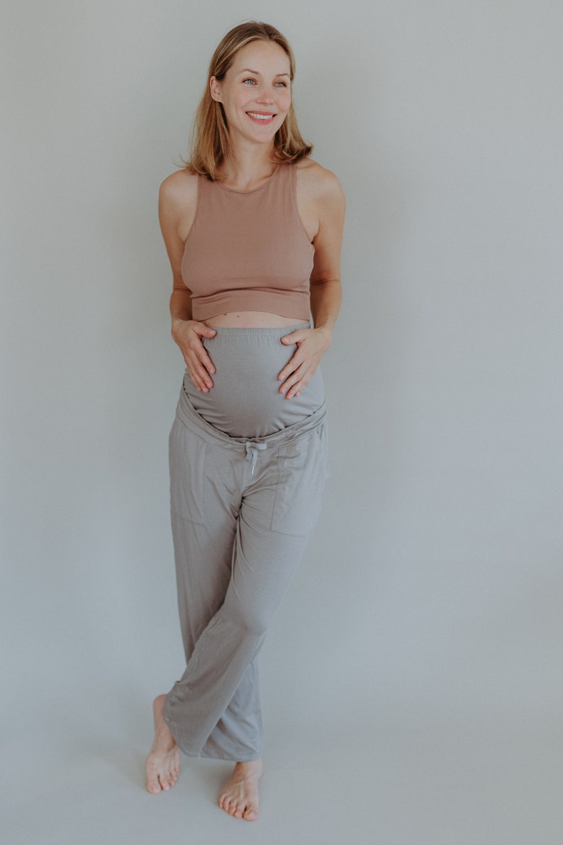 May include: A woman wearing a light brown tank top and gray maternity pants. The pants have a drawstring waist and pockets. The woman is smiling and holding her belly.