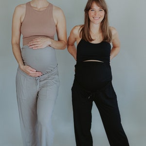 May include: Two women wearing maternity clothing. One woman wears a light brown tank top and gray pants. The other woman wears a black tank top and black pants. Both women are smiling and holding their bellies.