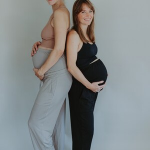May include: Two pregnant people stand back-to-back, wearing comfortable clothing. One wears a light brown tank top and gray pants, the other a black tank top and black pants. Both are smiling, with a neutral background.