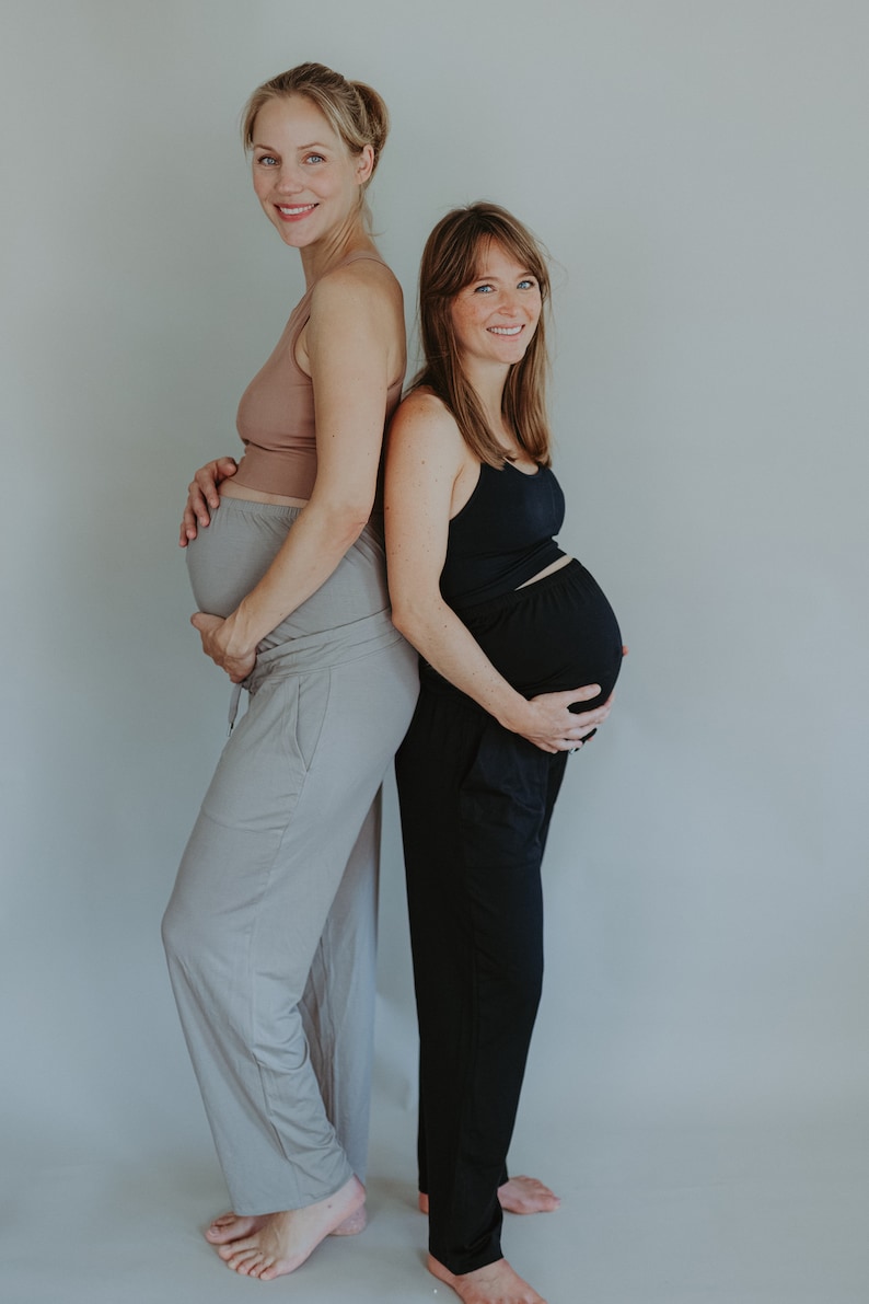 May include: Two pregnant people stand back-to-back, wearing comfortable clothing. One wears a light brown tank top and gray pants, the other a black tank top and black pants. Both are smiling, with a neutral background.