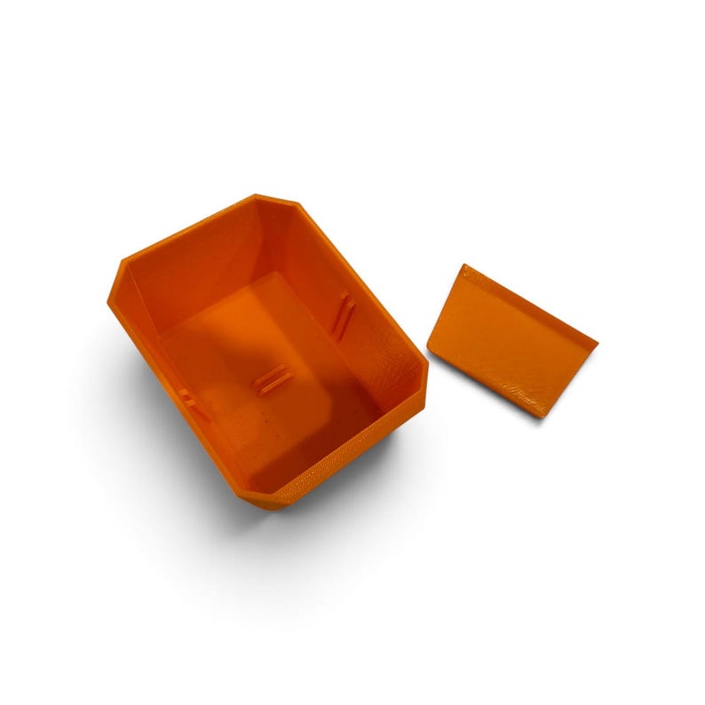 Op de afbeelding: Een feloranje opbergbak met open bovenkant, een rechthoekige basis en schuine zijden, inclusief een apart, bijpassend oranje rechthoekig deksel dat aan de rechterkant ligt. De binnenkant van de bak toont kleine structurele ribbels op de wanden.