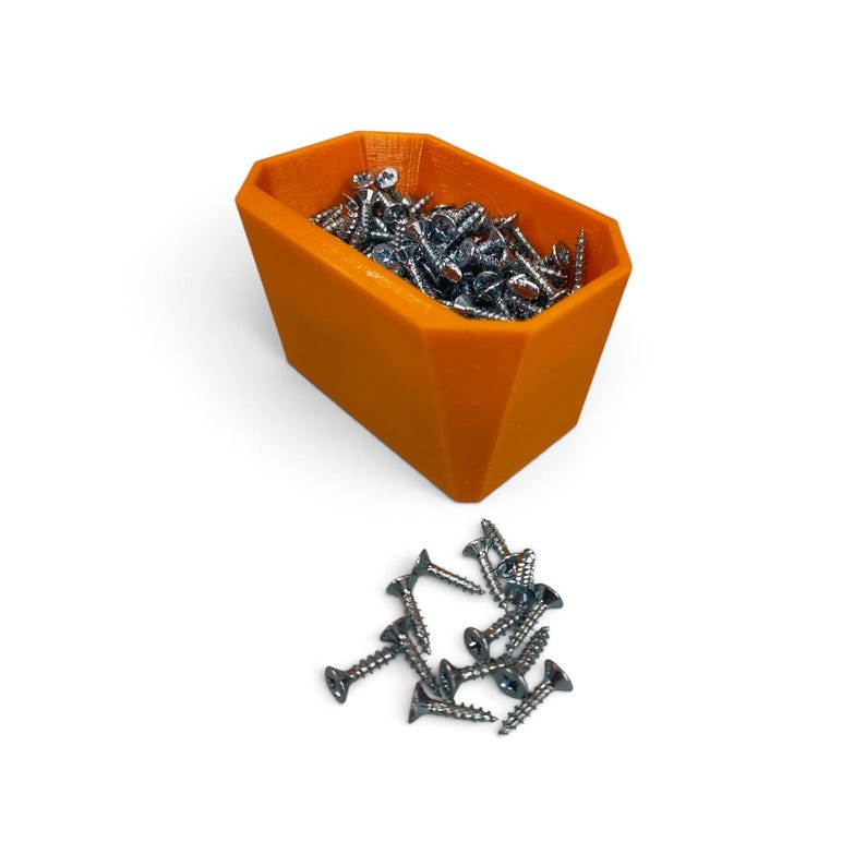 May include: A bright orange, faceted, open-top storage bin filled with numerous small, silver-colored metal screws. A small pile of identical silver metal screws is arranged on the surface directly in front of the orange container.