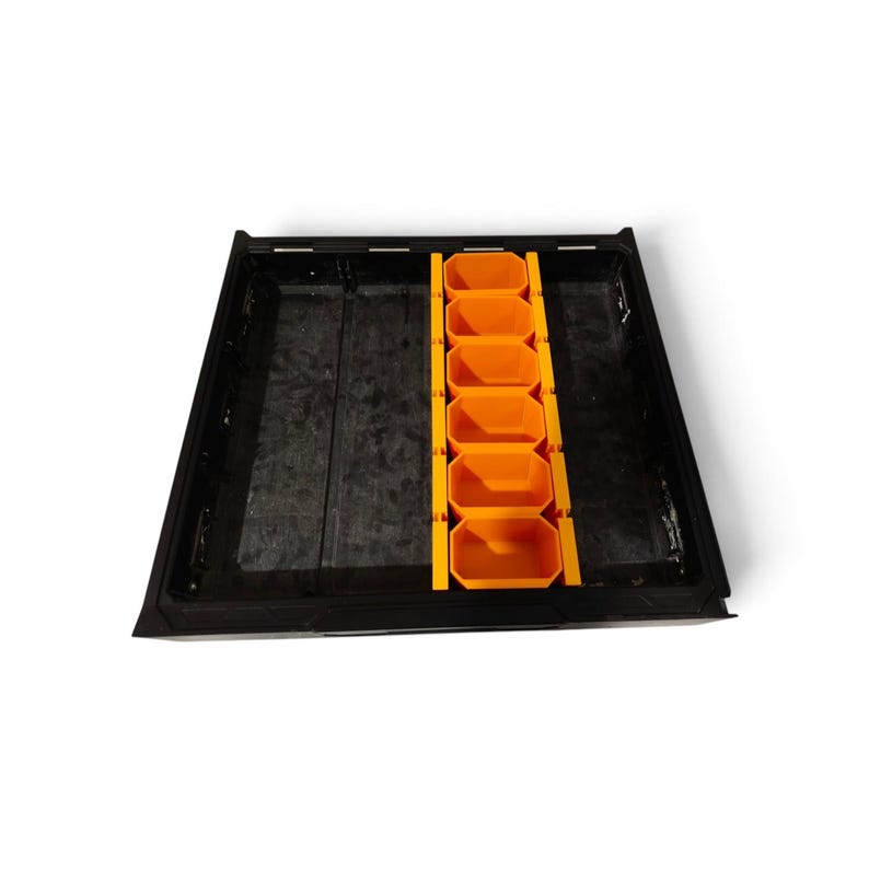 May include: A black rectangular storage tray featuring a central orange insert with six individual hexagonal compartments. The tray has a smooth, dark base and a structured frame, providing a partitioned space for organizing small items.