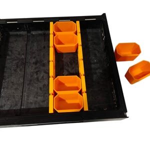 May include: A black rectangular storage tray featuring a central orange divider rail with multiple slots. Four orange bins are positioned within the tray, while two additional orange bins sit separately to the right of the main unit.