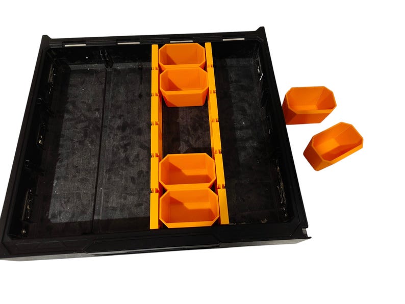 May include: A black rectangular storage tray featuring a central orange divider rail with multiple slots. Four orange bins are positioned within the tray, while two additional orange bins sit separately to the right of the main unit.