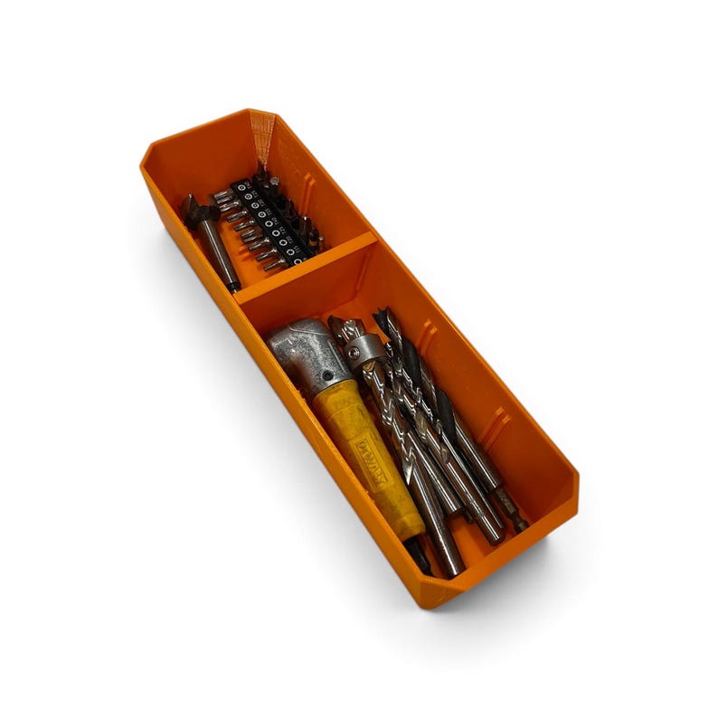 Op de afbeelding: Een oranje rechthoekige opbergbak met twee compartimenten. Het kleinere gedeelte bevat een rij metalen schroefbits. Het grotere gedeelte bevat een geel haaks boorhulpstuk en diverse metalen boortjes van verschillende afmetingen.
