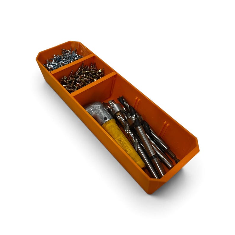 Op de afbeelding: Een oranje kunststof opbergbak met drie aparte vakken. De twee kleinere vakken bevatten zilver- en goudkleurige metalen schroeven, terwijl het grootste vak een gereedschap met een geel handvat en diverse metalen boortjes bevat.