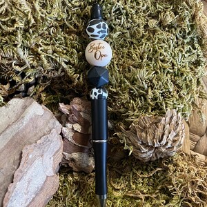 May include: A black pen with a wooden bead that says "Bester Opa" in gold lettering. The pen is decorated with black and white patterned beads.