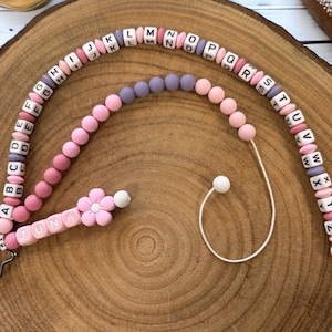 Może przedstawiać: Naszyjnik z koralików alfabetu w kolorze różowym, fioletowym i białym z zawieszką w kształcie kwiatu i imieniem "JUNA" napisanym różowymi koralikami. Naszyjnik jest przymocowany do białego sznurka z białym koralikiem na końcu.