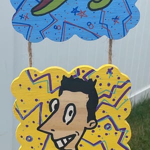May include: A whimsical, hand-painted wooden hanging decoration featuring three cartoon characters. The top panel shows a green pterodactyl on a blue background. The middle panel has a man with a bow tie on a yellow background. The bottom panel has a blue face on a red background.