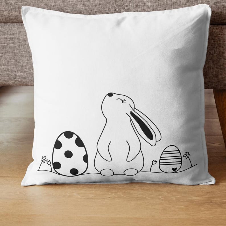 Pu&ograve; includere: Un cuscino bianco con un'illustrazione in bianco e nero di un coniglio che guarda in alto, con due uova di Pasqua su entrambi i lati. Un uovo &egrave; a pois e l'altro ha delle strisce.