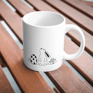 Pu&ograve; includere: Tazza in ceramica bianca con un disegno a linea nera di un coniglio pasquale e uova. Il coniglio guarda in alto e ha le orecchie dritte. Le uova sono decorate con pois e linee.