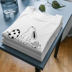 Pu&ograve; includere: Una maglietta bianca con un'illustrazione in bianco e nero di un coniglio e uova di Pasqua. Il coniglio sta guardando in alto e ha gli occhi chiusi. Le uova sono decorate con pois e strisce.