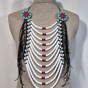 May include: A multi-strand beaded necklace featuring ten cascading rows of white, black, and turquoise beads. Two circular medallions with a red and turquoise star pattern anchor the strands, which are accented by long, thin black leather fringe.