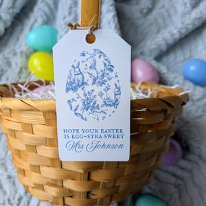 May include: A woven basket filled with Easter grass and colorful plastic eggs. A white tag with a blue floral egg design and the text "HOPE YOUR EASTER IS EGG-STRA SWEET Mrs Johnson" hangs from the basket.