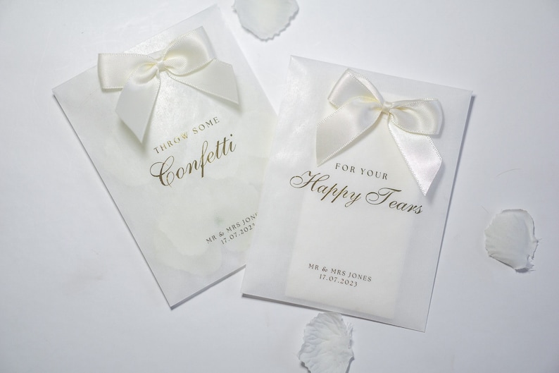 Pu&ograve; includere: Due sacchetti bianchi per bomboniere di matrimonio con scritte dorate. Un sacchetto dice "Throw some confetti" e l'altro dice "For your happy tears". Entrambi i sacchetti hanno fiocchi di raso bianco e sono personalizzati con i nomi "MR & MRS JONES" e la data "17.07.2023".