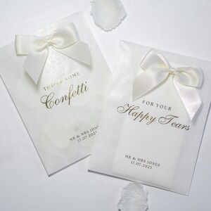 Pu&ograve; includere: Due sacchetti bianchi per bomboniere da matrimonio con scritte dorate. Un sacchetto dice "Throw some confetti" e l'altro dice "For your happy tears". Entrambi i sacchetti hanno nastri di raso bianco legati a fiocco.