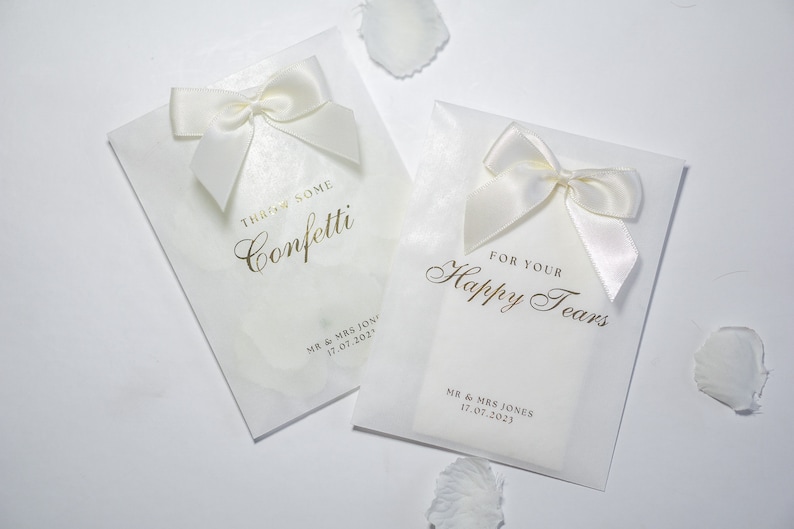 Pu&ograve; includere: Due sacchetti bianchi per bomboniere da matrimonio con scritte dorate. Un sacchetto dice "Throw some confetti" e l'altro dice "For your happy tears". Entrambi i sacchetti hanno nastri di raso bianco legati a fiocco.