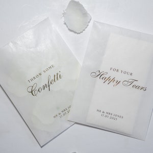 Pu&ograve; includere: Due sacchetti di carta bianca con lettere dorate. Un sacchetto dice "Throw some confetti" e l'altro dice "For your happy tears". Entrambi i sacchetti hanno la data "17.07.20" stampata.