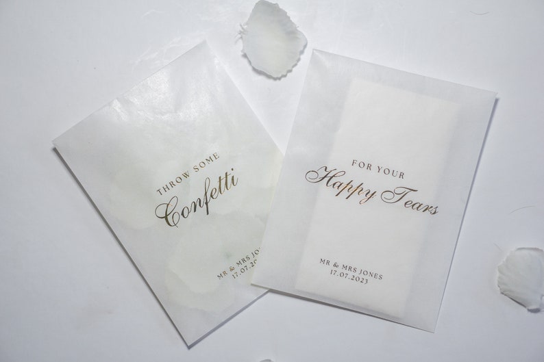 Pu&ograve; includere: Due sacchetti di carta bianca con lettere dorate. Un sacchetto dice "Throw some confetti" e l'altro dice "For your happy tears". Entrambi i sacchetti hanno la data "17.07.20" stampata.