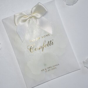 Pu&ograve; includere: Busta di carta bianca per confetti con un fiocco di raso bianco e scritta dorata che dice "Throw Some Confetti" e "Mr & Mrs Jones 17.07.2023".