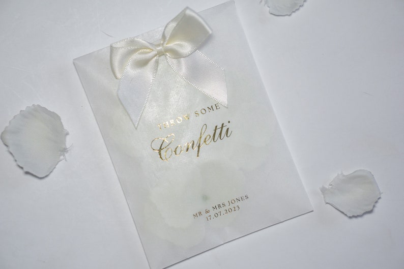Pu&ograve; includere: Busta di carta bianca per confetti con un fiocco di raso bianco e scritta dorata che dice "Throw Some Confetti" e "Mr & Mrs Jones 17.07.2023".