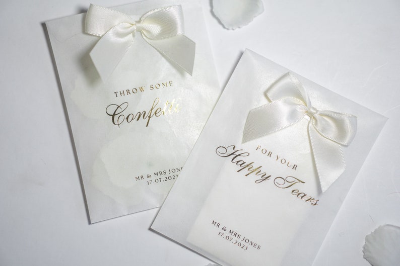 Pu&ograve; includere: Due sacchetti di carta bianchi con lettere dorate e fiocchi di raso bianco. Un sacchetto dice "Throw some confetti" e l'altro dice "For your happy tears". Entrambi i sacchetti hanno il testo "MR & MRS JONES 17.07.2023".