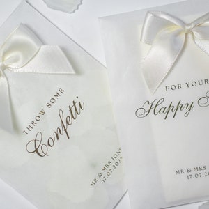 Pu&ograve; includere: Due sacchetti di carta bianca con fiocchi di raso bianco. Un sacchetto dice "Throw some confetti" e l'altro dice "For your happy tears". Entrambi i sacchetti hanno la data "17.07.2023" stampata.