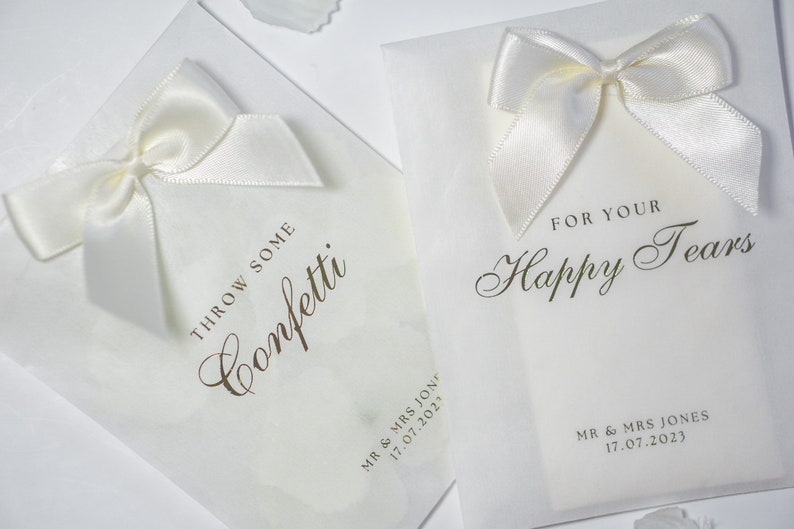 Pu&ograve; includere: Due sacchetti di carta bianca con fiocchi di raso bianco. Un sacchetto dice "Throw some confetti" e l'altro dice "For your happy tears". Entrambi i sacchetti hanno la data "17.07.2023" stampata.