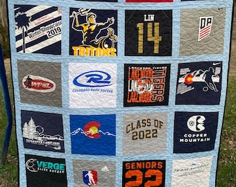 May include: A handmade quilt featuring squares of various t-shirts. The quilt showcases a variety of logos, text, and graphics, including sports teams, events, and school names. The quilt is bordered with a light blue and white checkered pattern.