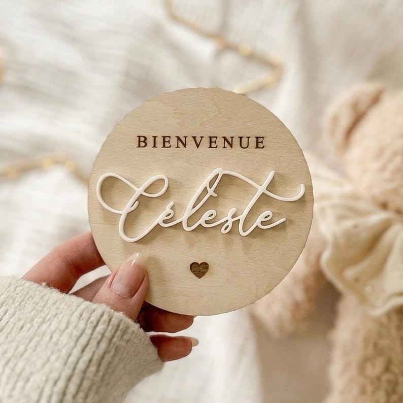 May include: A round wooden sign with the word "BIENVENUE" at the top and the name "Celeste" in cursive script. A small heart is carved at the bottom. The sign is held in a hand.