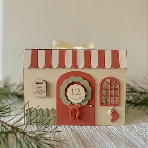 Peut inclure: Une maison en bois sur le thème de Noël avec un auvent à rayures rouges et blanches. La maison comprend une porte rouge avec une couronne verte et le chiffre 12, une fenêtre et une boîte aux lettres. Une petite chaussette est suspendue à la fenêtre. Le texte "Lettre au Père Noël" est visible.