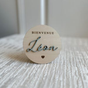 May include: A round wooden sign with the word "BIENVENUE" at the top and the name "L&eacute;on" in a cursive font. A small heart is below the name. The text and name are in a light blue color.