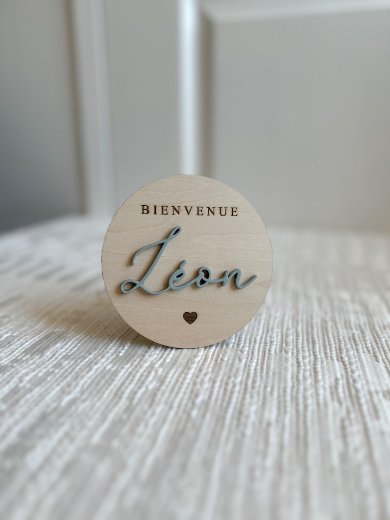 May include: A round wooden sign with the word "BIENVENUE" at the top and the name "L&eacute;on" in a cursive font. A small heart is below the name. The text and name are in a light blue color.