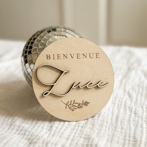 May include: A round wooden sign with the word "BIENVENUE" at the top and the name "Luce" in cursive script. A small floral design is below the name. The sign is light brown and rests in front of a disco ball.