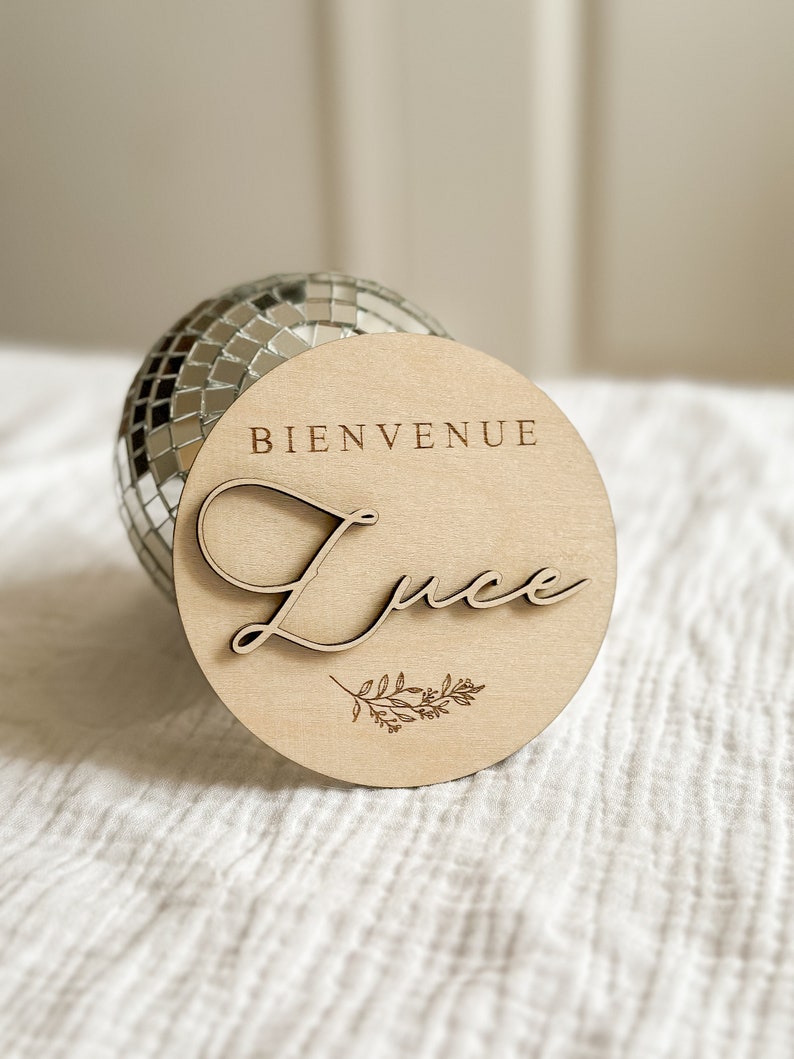 May include: A round wooden sign with the word "BIENVENUE" at the top and the name "Luce" in cursive script. A small floral design is below the name. The sign is light brown and rests in front of a disco ball.